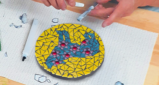 Une personne place soigneusement des carreaux de verre colorés sur un panneau de mosaïque circulaire représentant un loup de couleur turquoise sur un fond jaune.