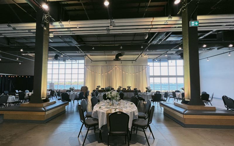 Un ensemble pour banquet de mariage et des tables ovales et une table d’honneur placées entre deux fenêtres allant du sol au plafond et donnant sur le port. La table se trouvant derrière le mur d’honneur est décorée de rideaux blancs et de lumières.