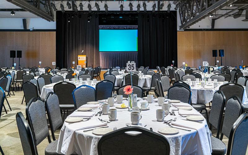 Un espace prévu pour un banquet offrant des tables ovales et des chaises de banquet. Chaque table présente un panneau de numéro de table et un vase-bouteille contenant une marguerite. On y trouve une scène offrant un podium, un microphone et une toile de projection.