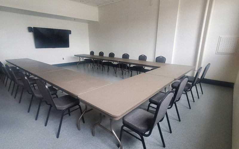 Une petite pièce aux hauts plafonds pouvant accueillir quinze invités, équipée de tables rectangulaires et de chaises de banquet disposées en forme de U.  L’installation fait face à un téléviseur en vue d’une présentation.