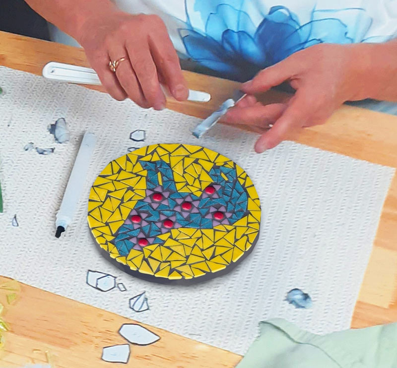 Une personne place soigneusement des carreaux de verre colorés sur un panneau de mosaïque circulaire représentant un loup de couleur turquoise sur un fond jaune.