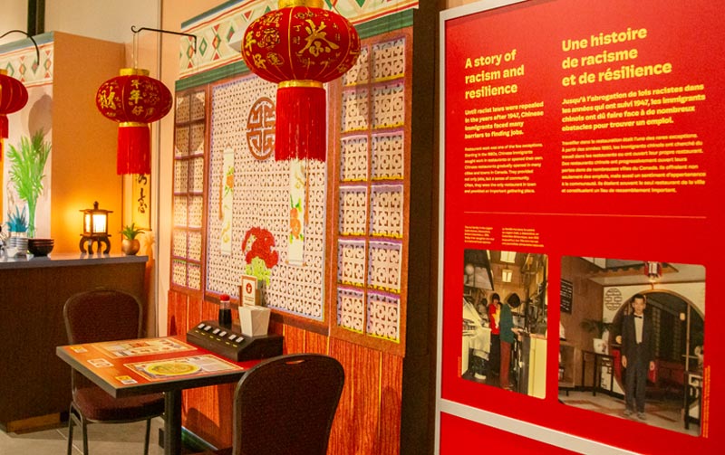 Aire d’exposition qui ressemble à un restaurant sino-canadien, avec une petite table, deux chaises et des lanternes rouges.