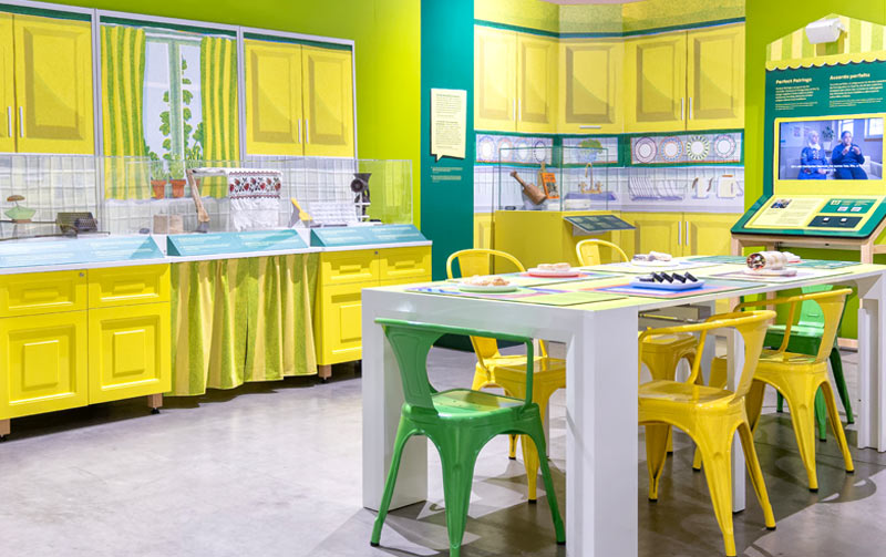 Aire d’exposition conçue pour ressembler à une cuisine, avec une table à manger, des chaises et des vitrines.