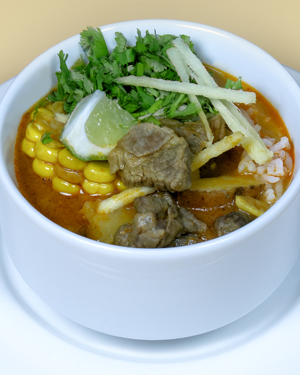 Un bol d'aloo birria avec un bouillon rougeâtre, des épis de maïs, des morceaux de bœuf, du riz, de la coriandre, des bandes de tortilla et un quartier de citron vert.