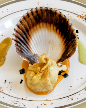 Un momo aux fruits de mer placé devant une coquille Saint-Jacques dans une assiette blanche, sur un fond de sauce orange, avec une sauce ail et coriandre à gauche et un chutney de timut à droite.