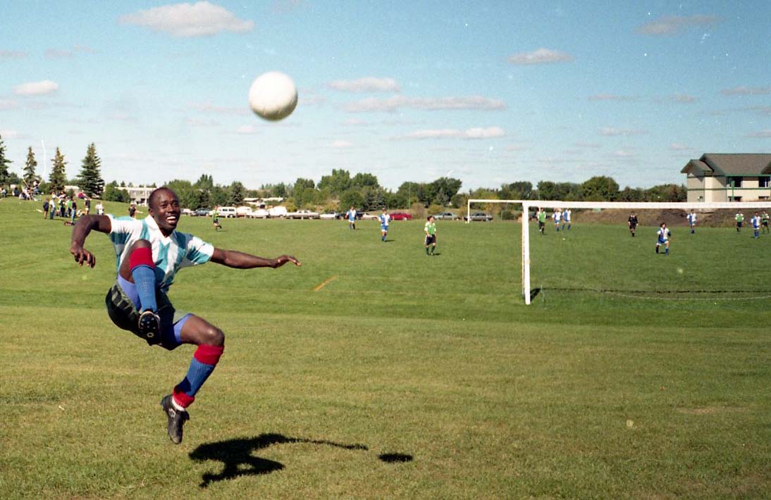 Un homme noir dont les pieds ne touchent plus le sol sourit après avoir botté un ballon. Il est en plein soleil, sur un grand terrain, devant terrain de soccer en activité.