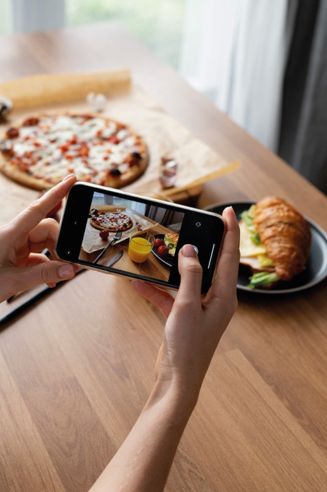 Une personne tient un téléphone intelligent et prend une photo d’un repas sur une table en bois, le repas est constitué d’une pizza, d’un croissant sur une assiette, d’un verre de jus d’orange et de tomates cerises.