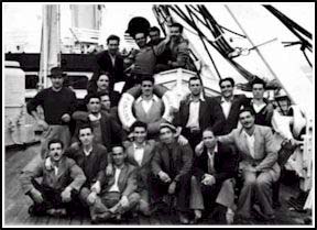 Grand groupe de jeunes hommes, certains assis dans un canot de sauvetage et d’autres se tenant autour, à bord d’un bateau.