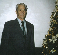 Un vieil homme en costume et cravate à côté d’un sapin.