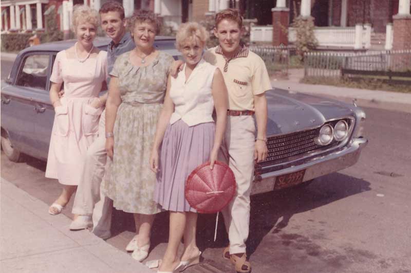 Trois femmes et deux hommes appuyés contre une vieille voiture bleue.