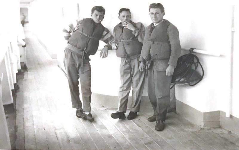 Trois hommes portant des gilets de sauvetage fument la cigarette sur le pont d’un navire.