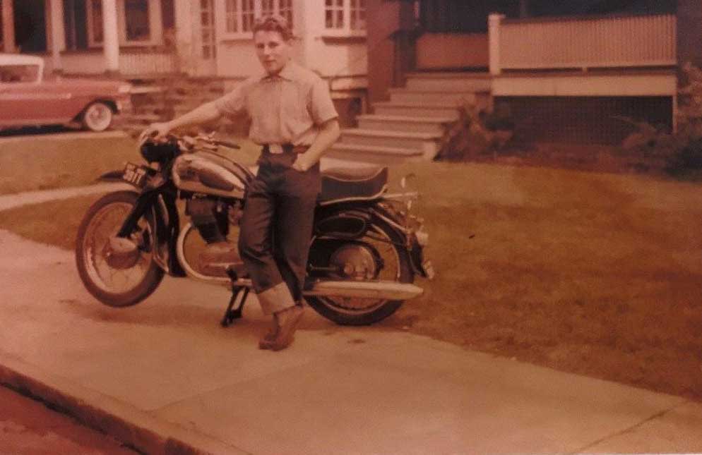 Un jeune homme appuyé contre une moto. L’une de ses mains est sur le guidon et l’autre dans sa poche.