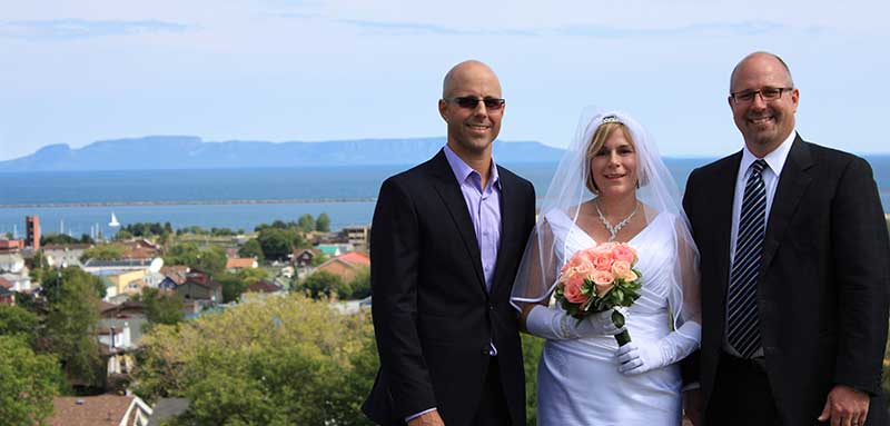 Une jeune épouse entre deux hommes bien habillés. Elle porte un voile et une robe et elle tient un bouquet de roses aux couleurs corail.