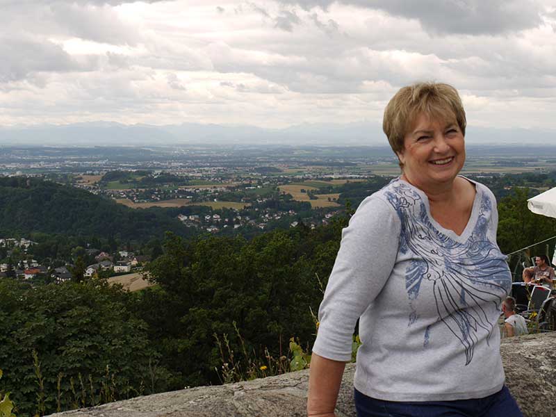 Photo récente d’une femme adossée à un mur de briques, et de beaux paysages derrière elle.