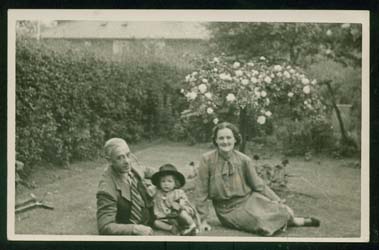 La jeune Joan portant un chapeau, assise dans l’herbe d’un jardin avec ses parents.