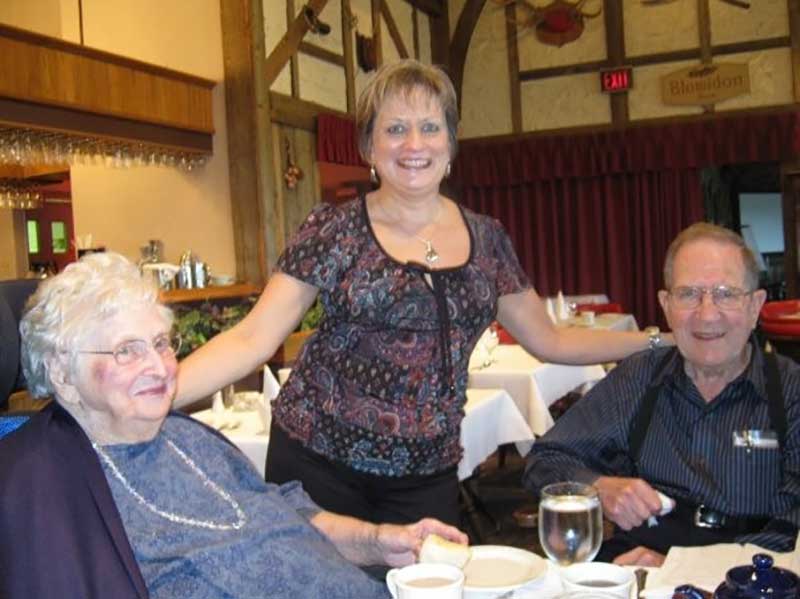 Un couple assis à une table. Une femme est debout entre le couple et a une main sur leurs épaules.
