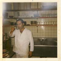 Nikolas en tenue de cuisinier blanche, debout devant un évier et des ustensiles de cuisine.