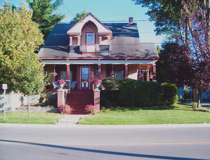 Une jolie maison de couleur corail. La photo a été prise de l’autre côté de la rue. La maison est entourée de plusieurs fleurs et d’une haie.