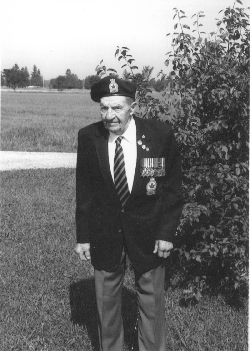 Norman plus âgé, portant l’uniforme de la légion, assis sur l’herbe devant un buisson.