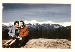 Un homme et une femme italiens assis sur une corniche rocheuse avec des arbres et des montagnes enneigées derrière eux.