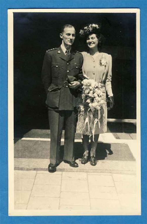 Mariée et marié le jour de leur mariage, le marié porte un uniforme militaire et la mariée nous tenant des fleurs et portant un grand chapeau.