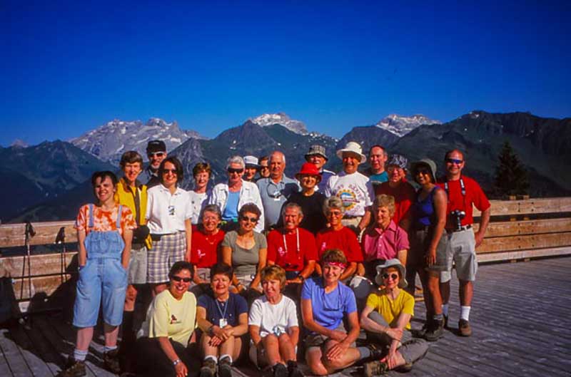 Un groupe de touristes se tiennent et s’agenouillent devant une paroi rocheuse, il y a de belles montagnes derrière eux.