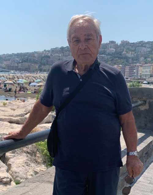 Un homme plus âgé dans un t-shirt bleu se tient contre une balustrade, il y a un fond scénique derrière lui.