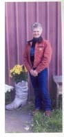 Photographie couleur d’une femme debout à côté d’un pot de fleurs en forme de botte.