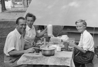 Photo en noir et blanc de trois personnes qui mangent à une table de pique-nique lors d’une sortie de camping.